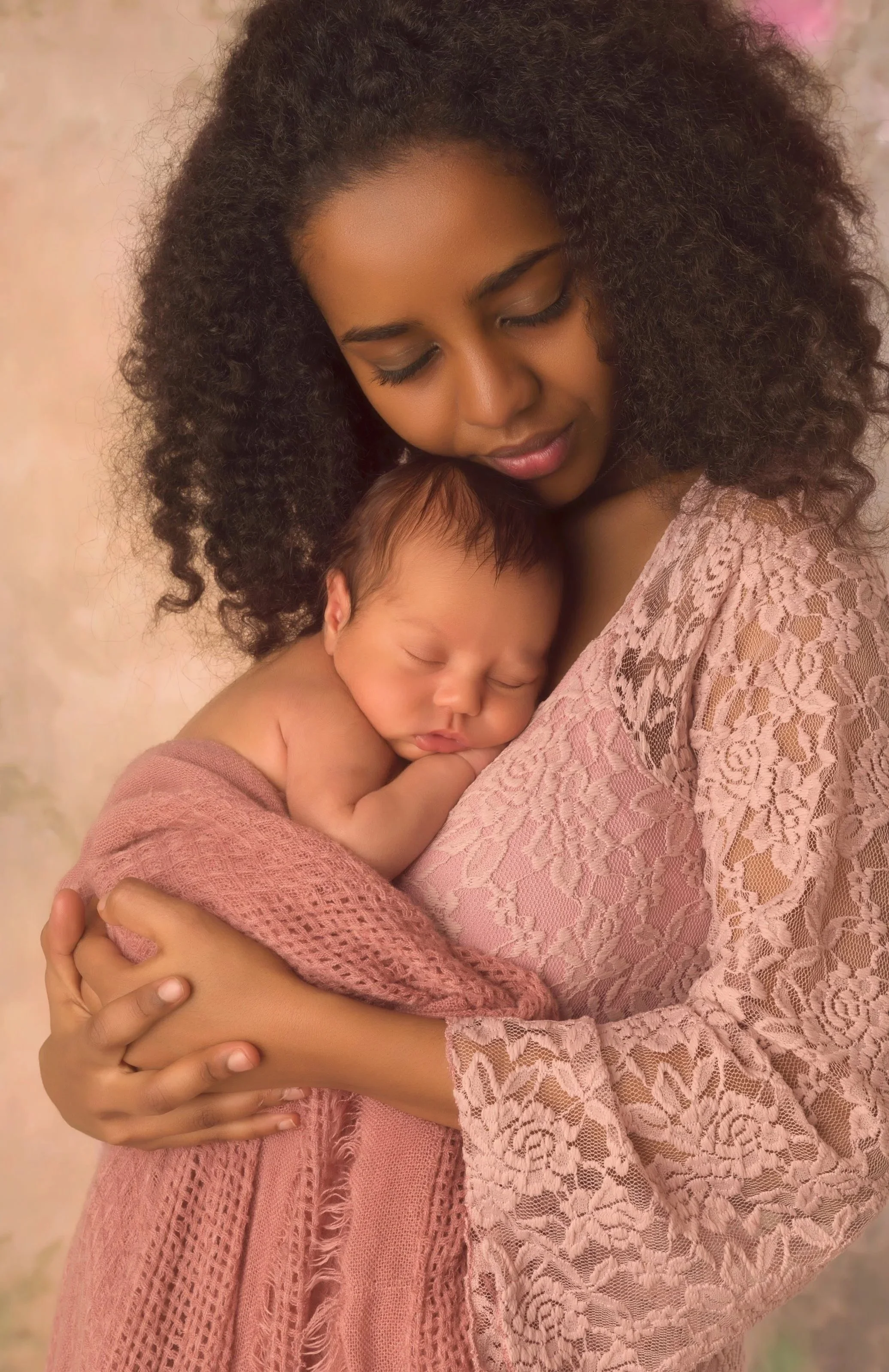 A woman with curly black hair holding a sleeping baby with light brown hair against her chest. The woman is wearing a pink lace top and the baby has a pink blanket wrapped around them.