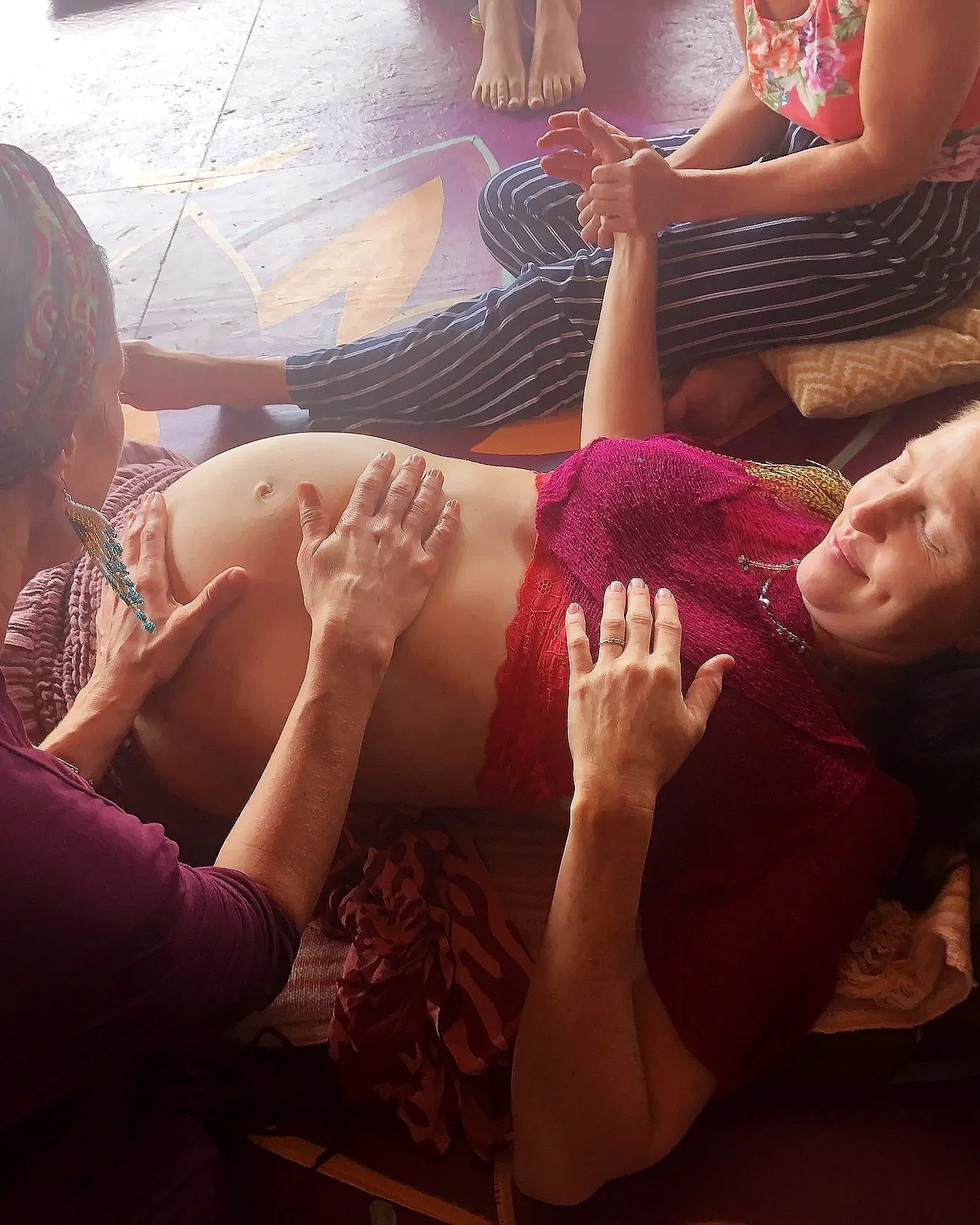 Group of women participating in an Ayurvedic massage session, with one woman lying face up and others around her, some touching her abdomen and others sitting nearby.