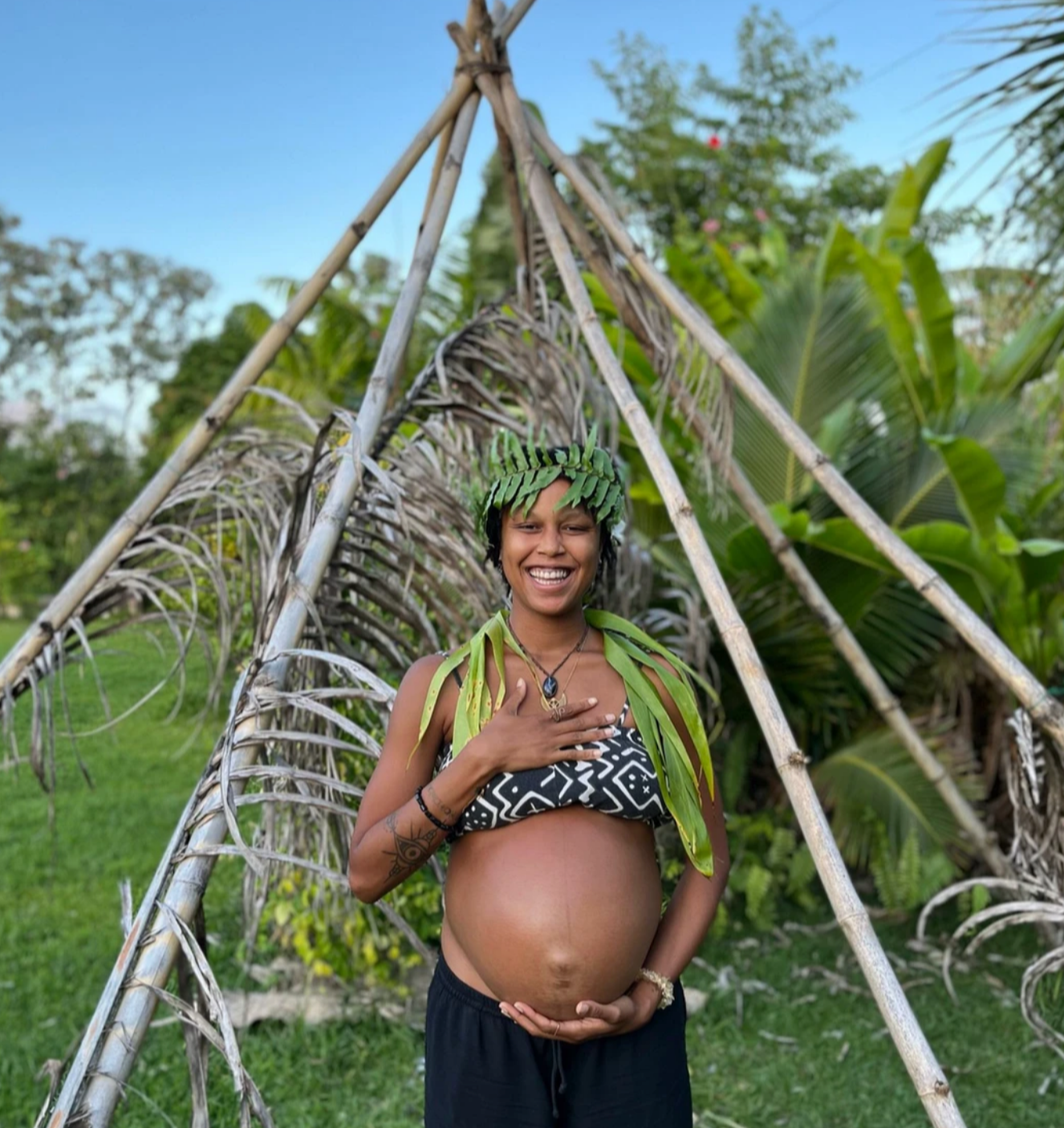 A joyful pregnant African American woman with a hand on her chest and the other on her belly, surrounded by lush tropical greenery in Hawaii during a Sacred Birth Path training with Hope Medford. SacredBirthPath.com