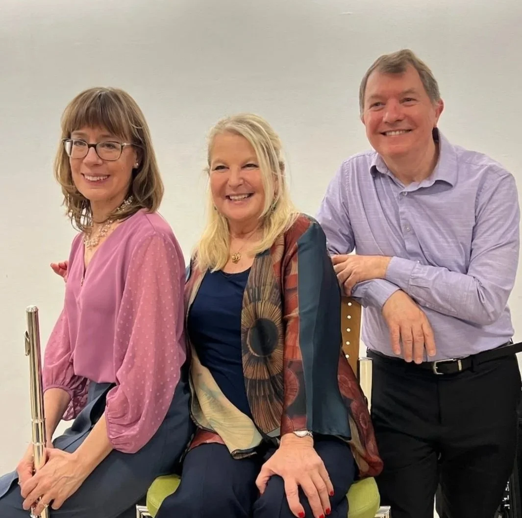 Three smiling individuals, two women and one man, pose together in a casual setting. The woman on the left holds a flute, the woman in the middle is seated, and the man on the right stands leaning on a chair. They are dressed in semi-formal attire.