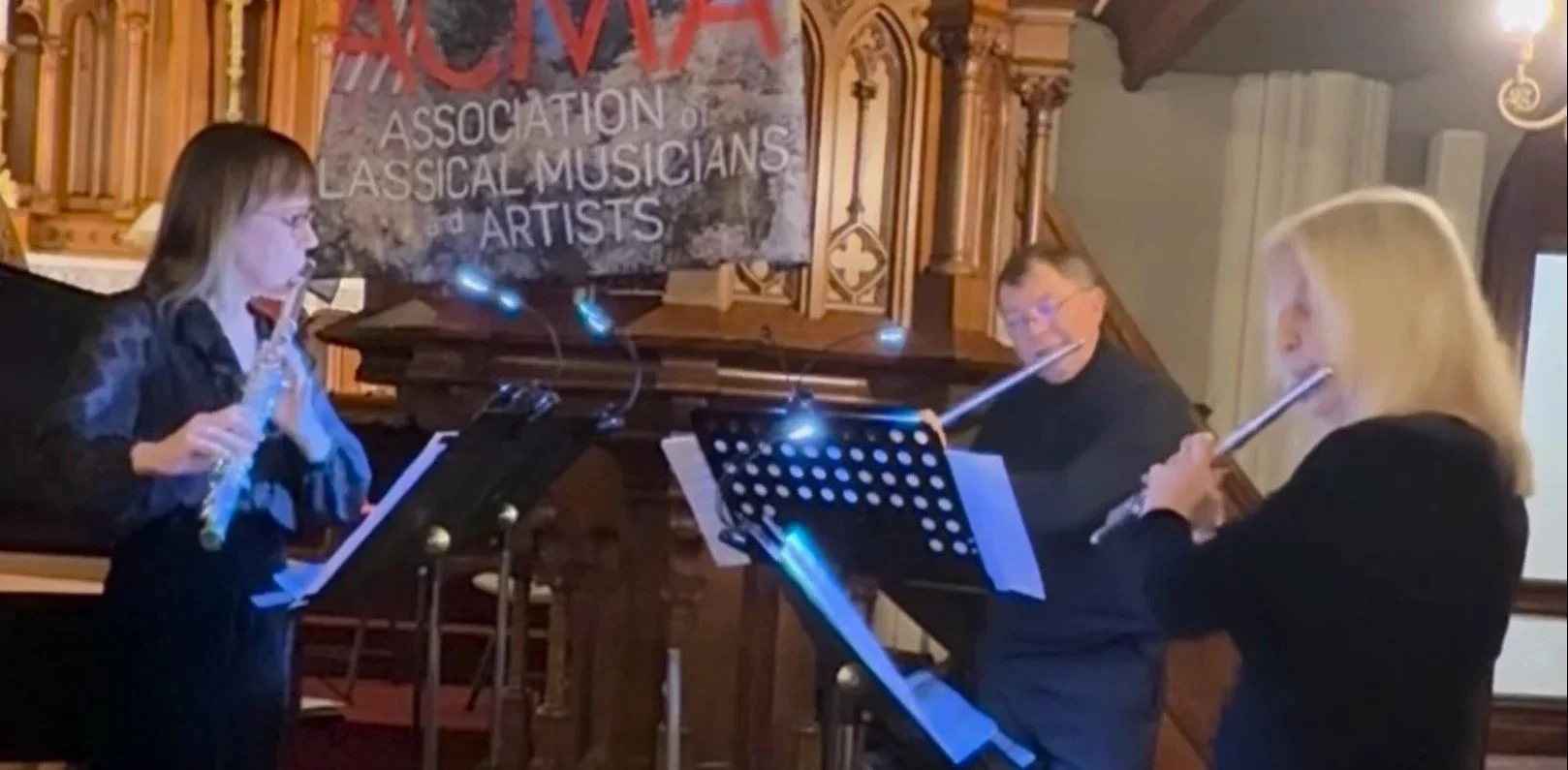 Three women playing flutes during a musical performance at a church or concert hall. A sign on the wall behind them reads 'CVM' and 'Association of Classical Musicians and Artists.'