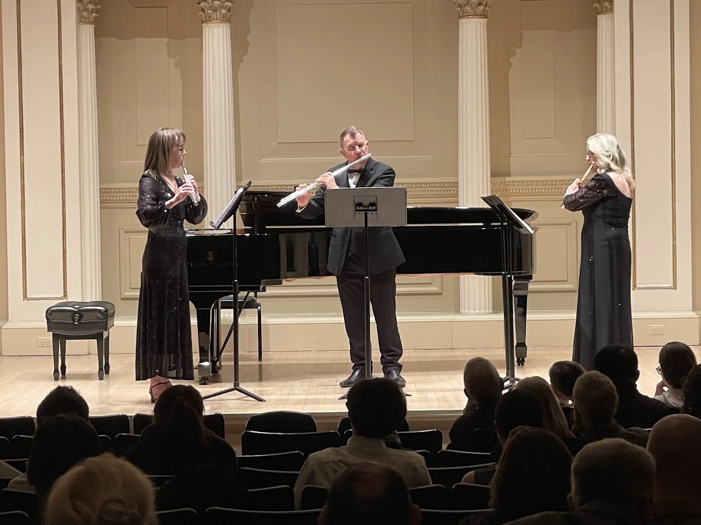 Three musicians performing on stage in front of an audience. A woman on the left plays the flute, a man in the center plays the flute, and a woman on the right plays the flute. They are dressed in formal attire, and there is a grand piano behind them