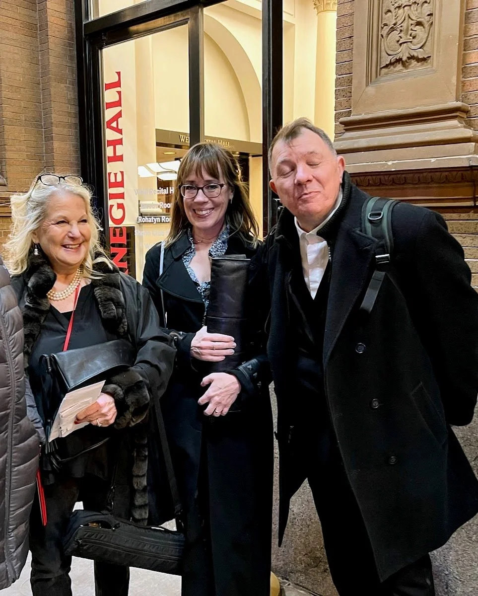 Three smiling people standing outside a hospital, with a sign for a regional hospital and a red sign reading 'Emergency Hall' visible behind them.