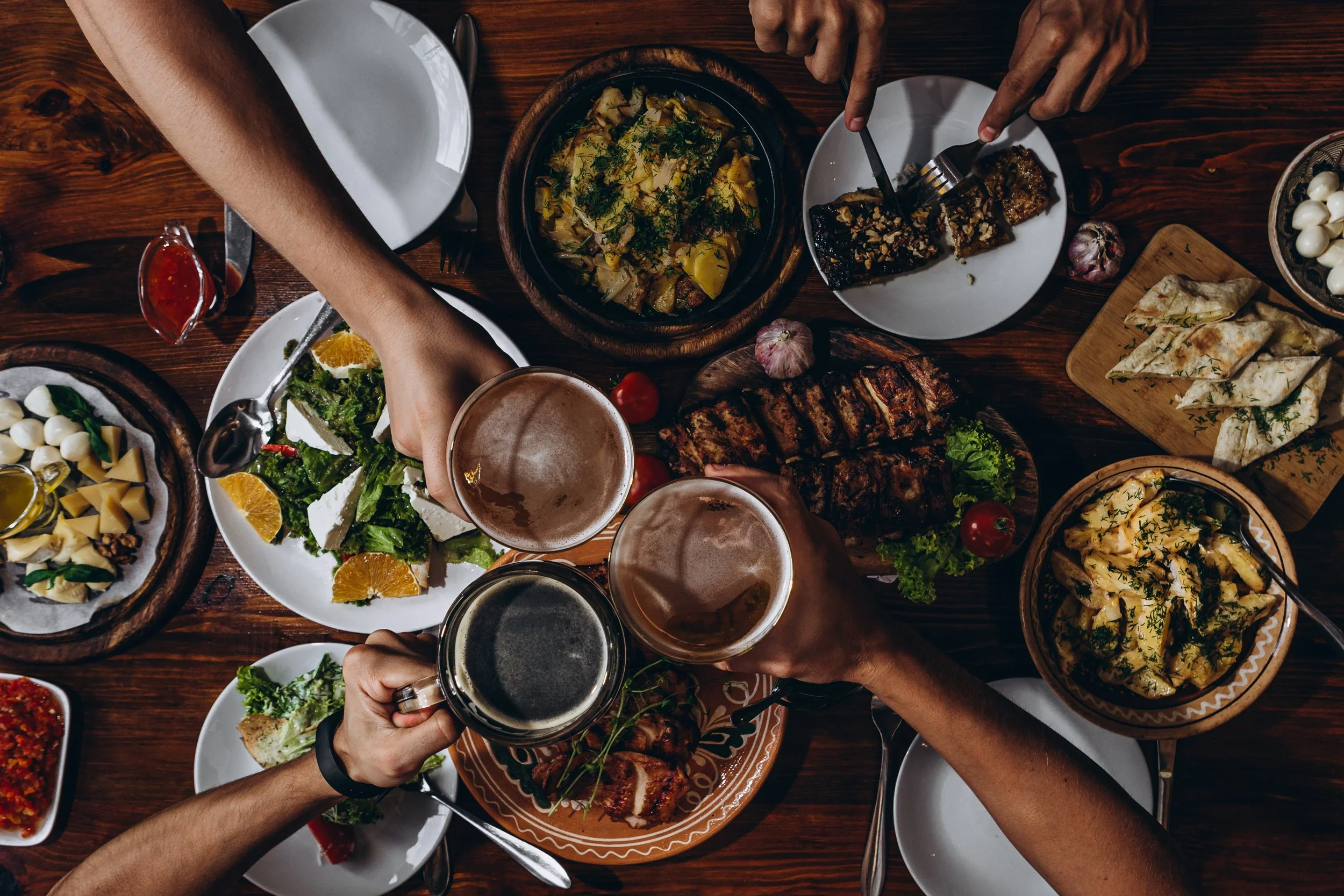 Top-down view of a dinner table with various dishes, three people raising glasses for a toast, including beer and wine, with plates of salad, grilled meat, bread, pasta, and sauces on a wooden table.
