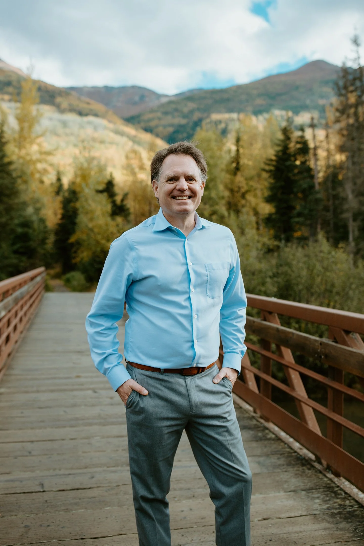 A man in a light blue dress shirt and gray pants standing on a wooden bridge with a scenic mountain and forest background.