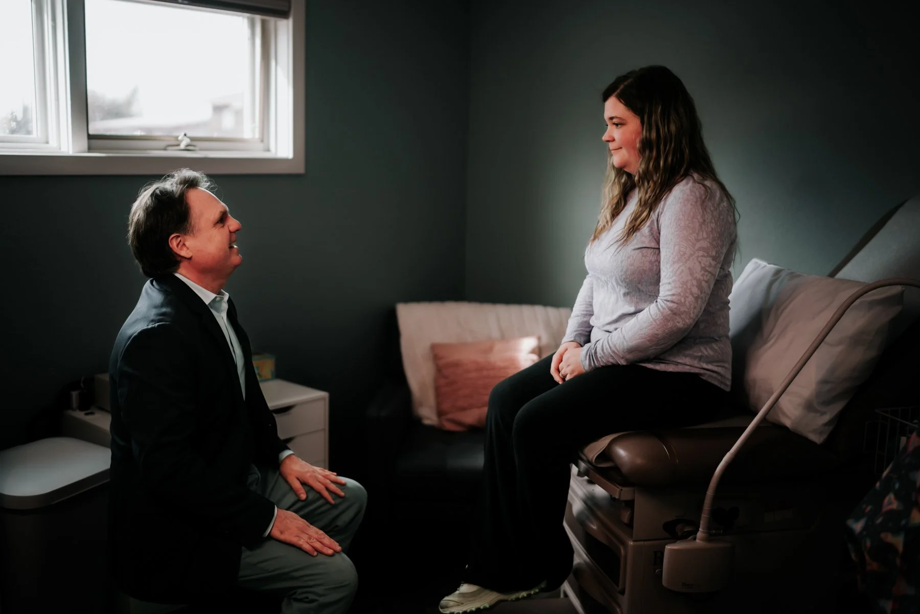 A woman sitting on an examination table in a doctor's office, smiling and talking to a man who is kneeling in front of her and smiling back.