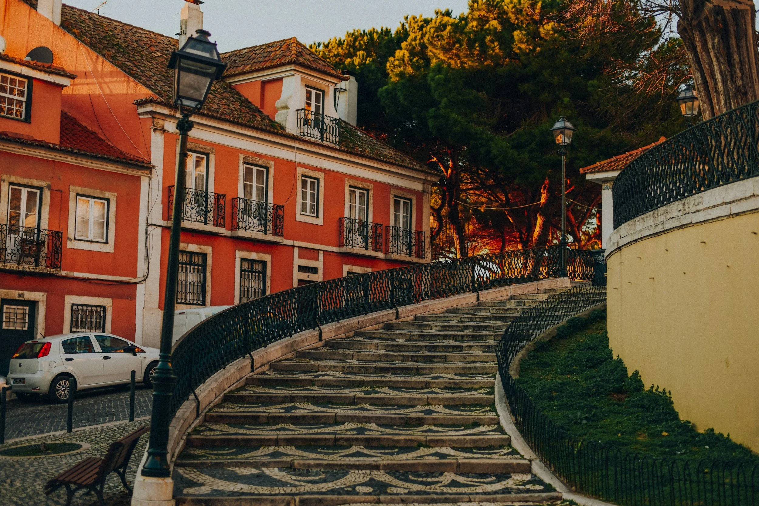 Colorful buildings with wrought iron balconies, a curved staircase with decorative paving, lampposts, parked cars, and trees in the background under a setting sun.