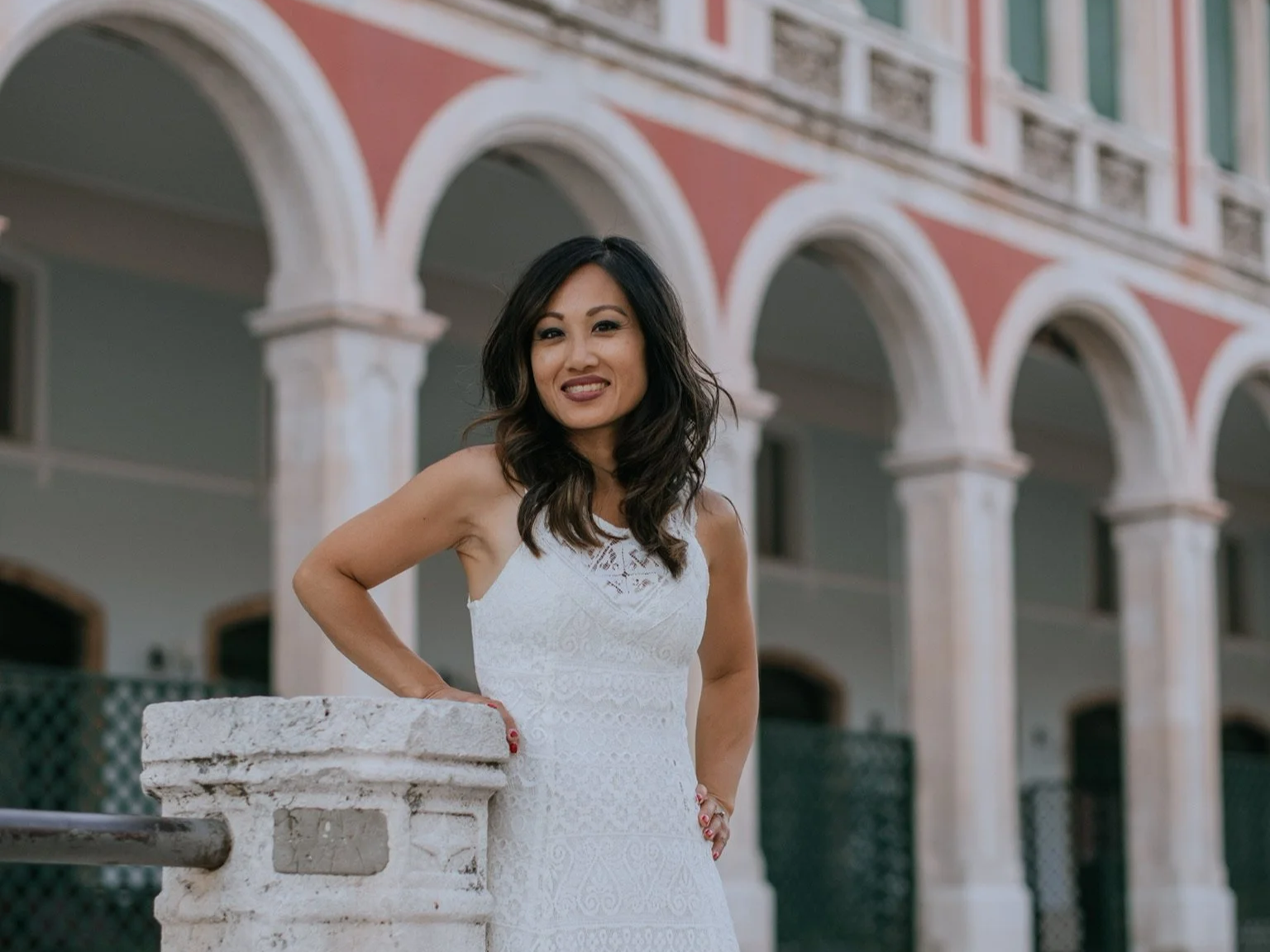 A woman in a white sleeveless dress leaning on a stone railing in front of a colorful building with arches.
