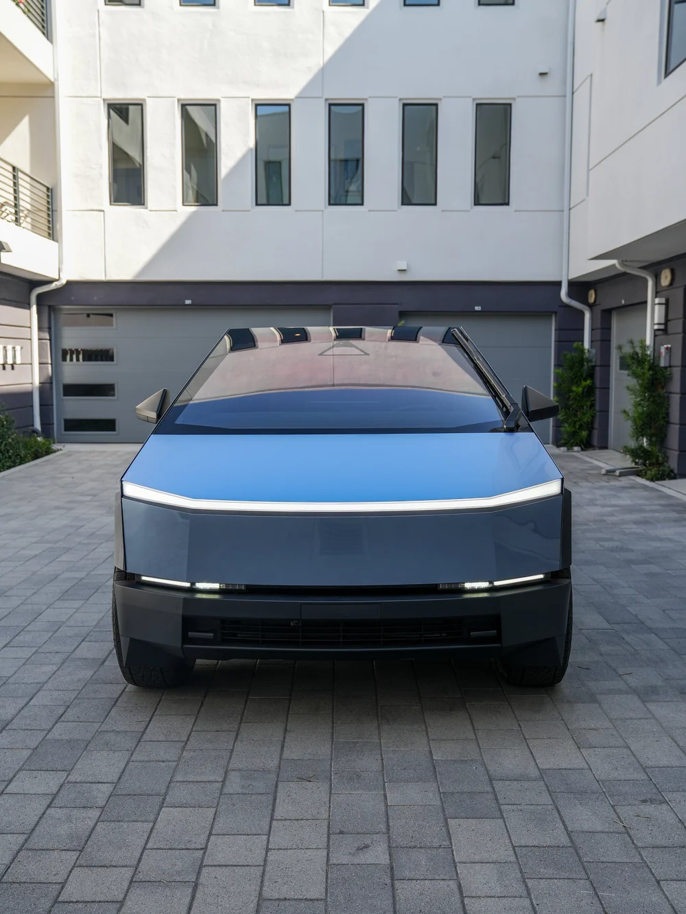 Straight front view of the blue-gray Cybertruck with a broad hood, white lamp strip, dark windshield and black lower bumper on courtyard paving.
