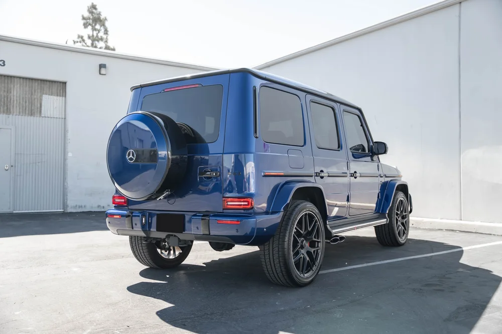 Complete opposite rear-three-quarter view of the blue G 63 with a reflective spare-wheel cover, dark wheels and twin side exhaust tips