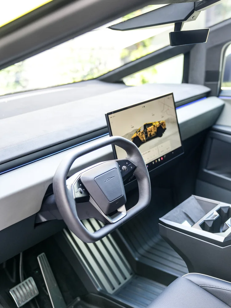 Tesla Cybertruck cockpit viewed through an open doorway, showing the steering wheel, central touchscreen, blue dashboard lighting, center console and pedals.