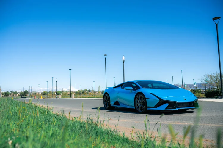 Blue Lamborghini Huracan Tecnica in a complete front three-quarter view beside grass, with gray wheels beneath a blue sky.