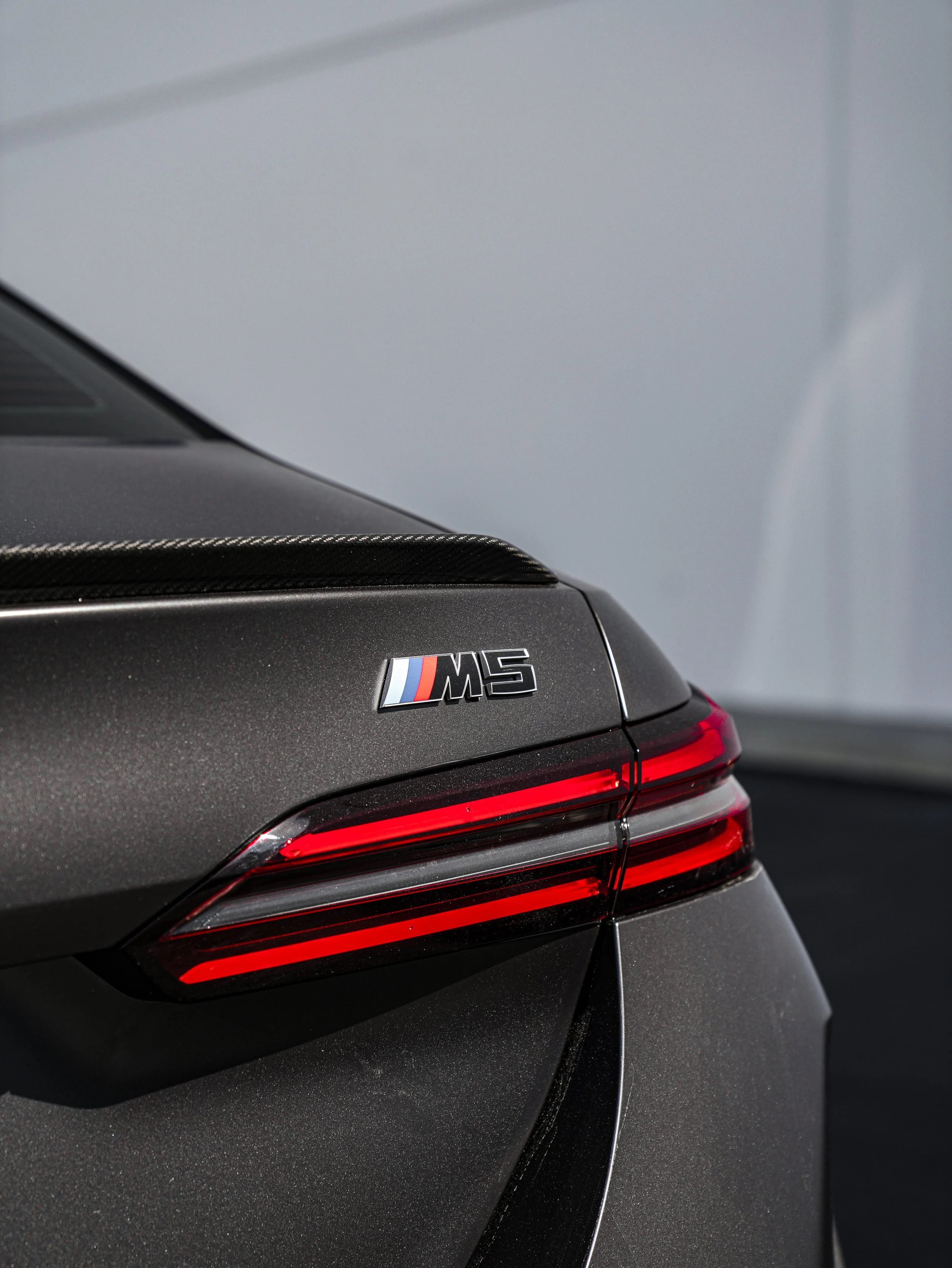 Close-up of the rear end of a dark gray BMW M5 with red tail lights and a carbon fiber trim on the trunk.