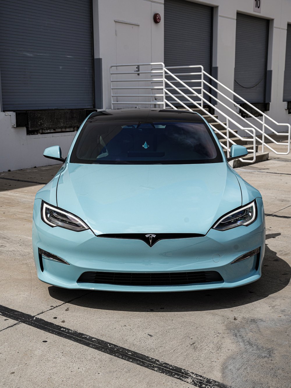 Pale blue Tesla Model S viewed directly from the front outside grey roller doors and white stairs, with both mirrors visible.