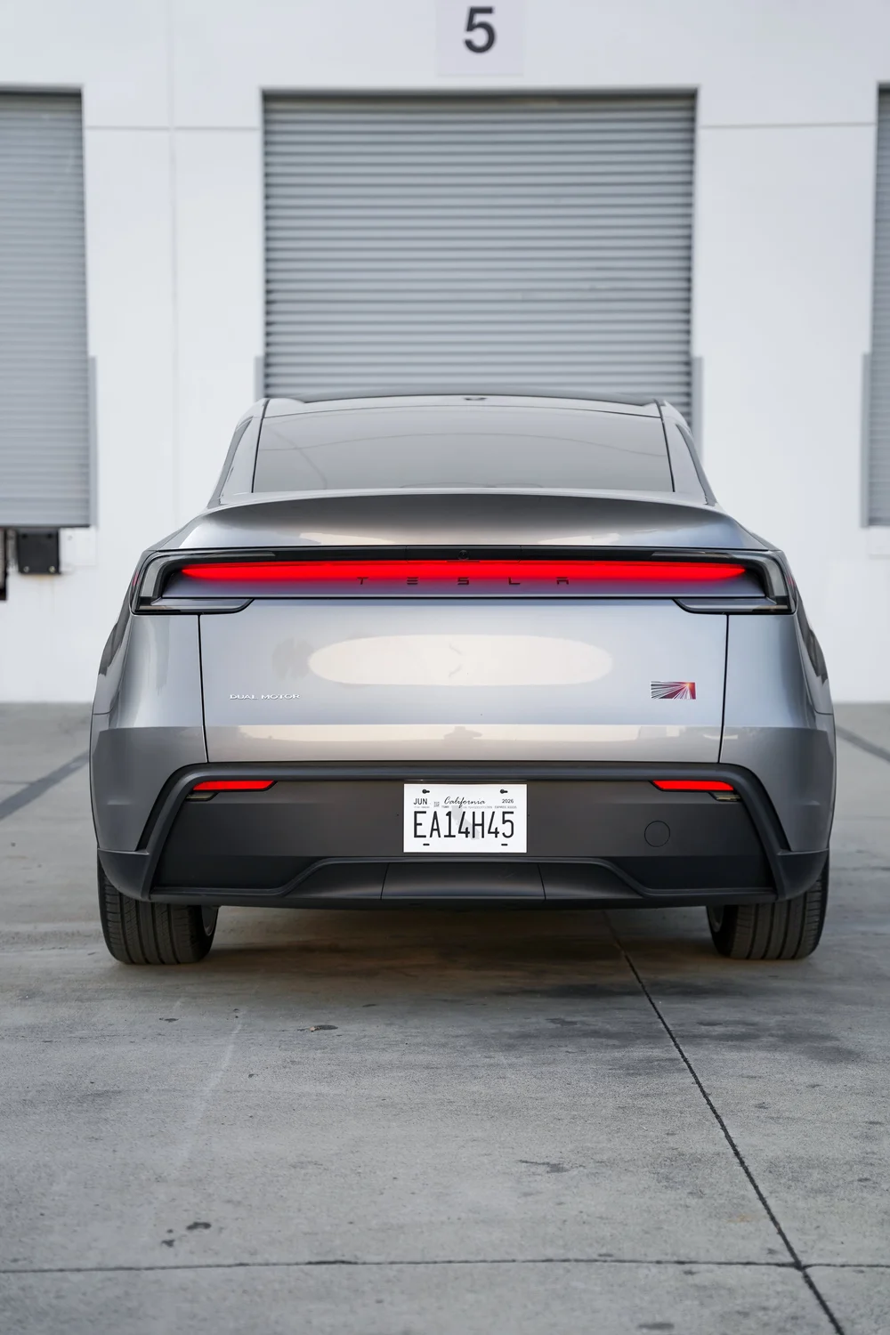 Complete straight rear view of the gray Tesla Model Y with a red light bar, liftgate, dark lower bumper and both rear tyres outside workshop doors.