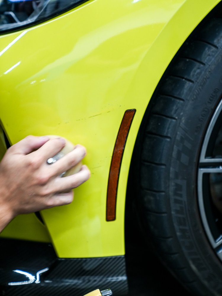 Hand holding a light cloth or pad against the yellow-green BMW M4 front-corner panel beside an amber reflector and front wheel inside the shop.