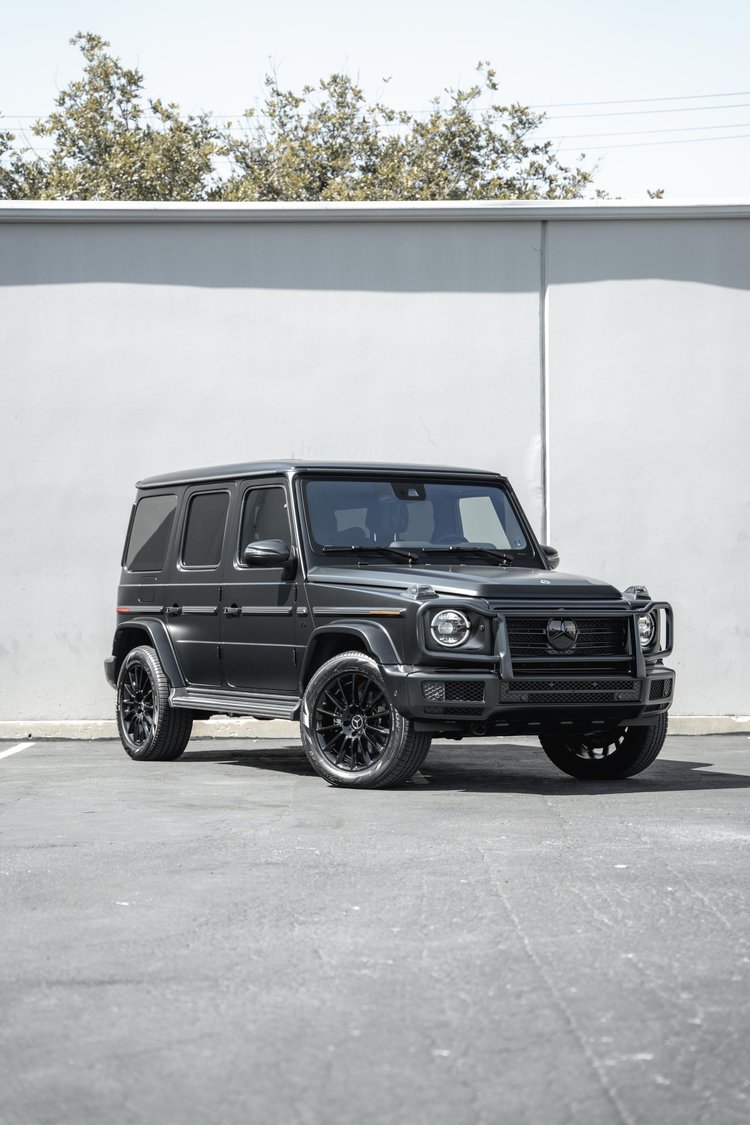 Tall complete front-three-quarter view of the grey G 550 with black wheels and a dark front guard below a white wall and trees