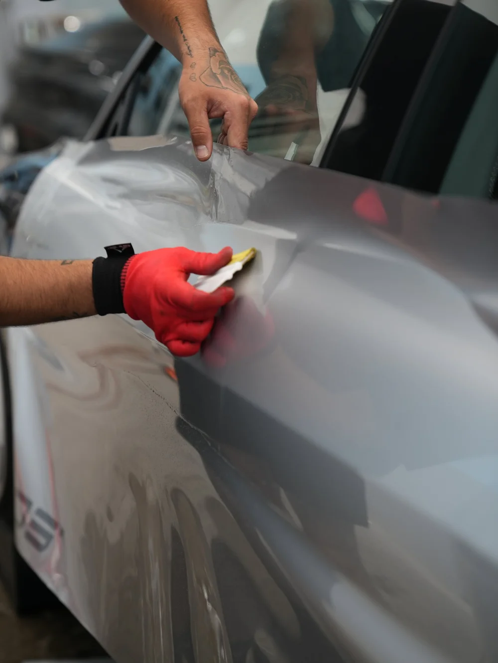 A red-gloved hand using a squeegee on silver film along a Model 3 door while another hand holds the loose film edge below the side window.