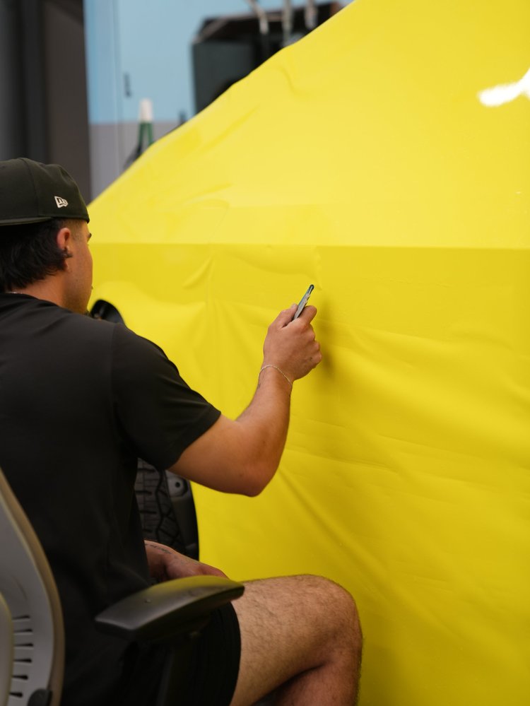 A technician holding a small hand tool beside a large loose sheet of yellow vinyl over an upright Tesla Cybertruck body panel.