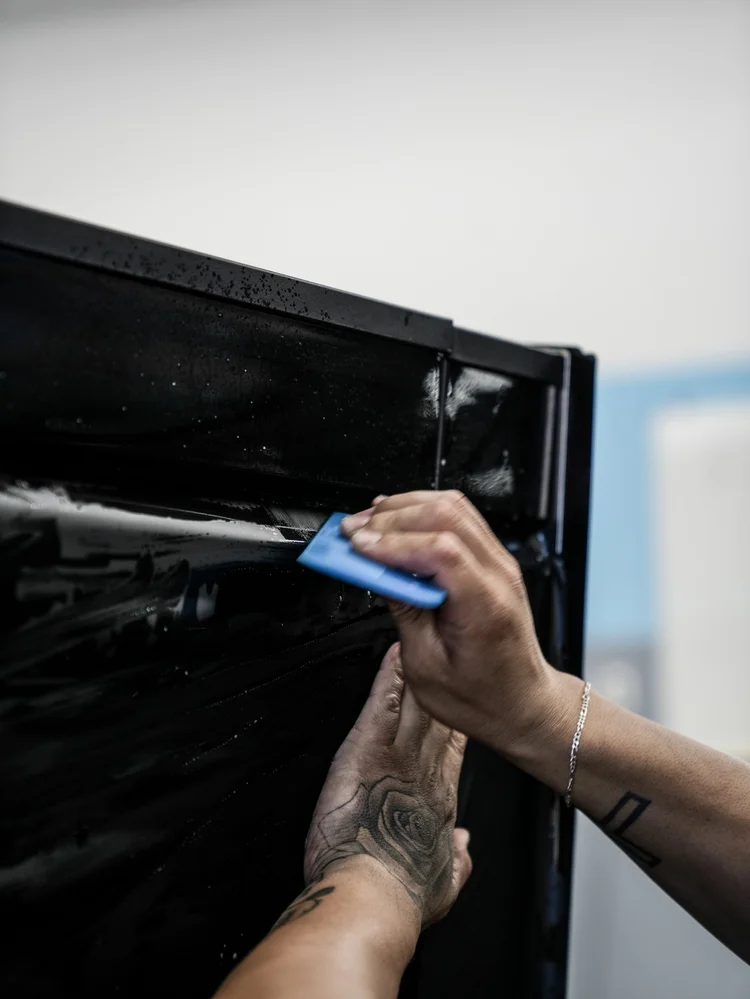 Hands pressing a blue squeegee along wet black film near the upper channel of an upright Tesla Cybertruck panel.