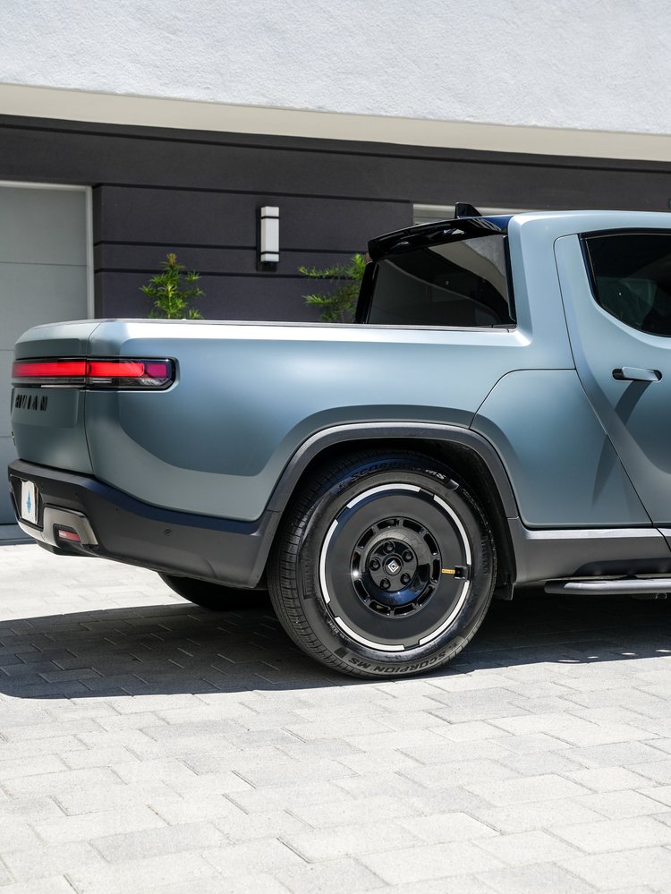 Rivian R1T rear-side detail showing satin bed-side panels, red tail lamp, black rear wheel, rear glass and side step.