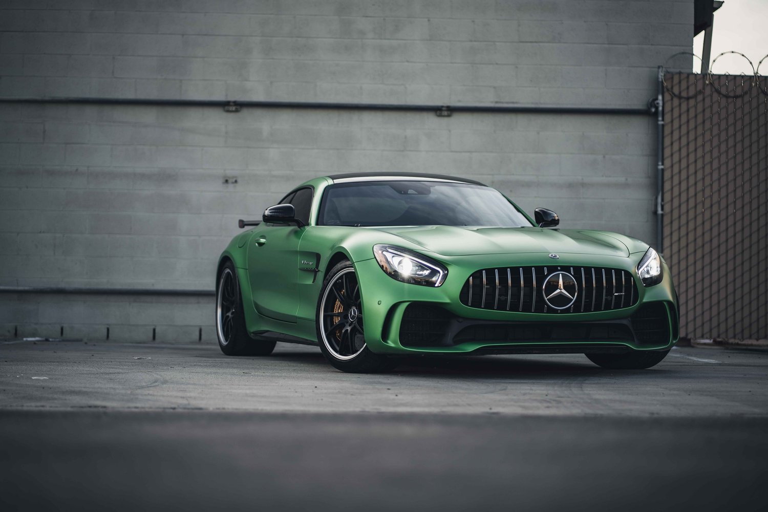 Complete green Mercedes-AMG GT R from a low front three-quarter angle, with illuminated lamps against a gray wall.