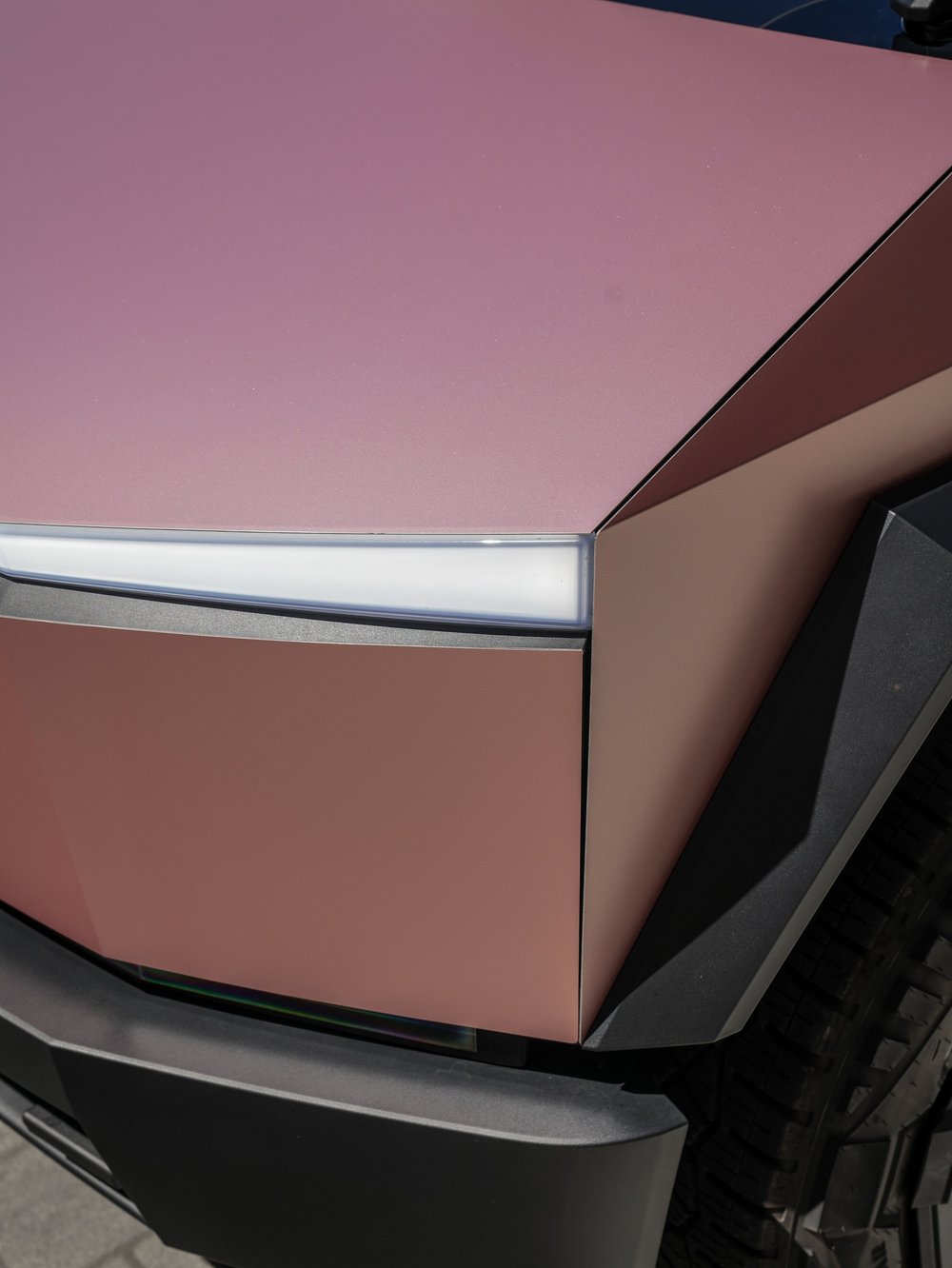 Close front corner of the Cybertruck showing pink-gold hood and fender surfaces beside the white lamp strip and dark wheel-arch trim.