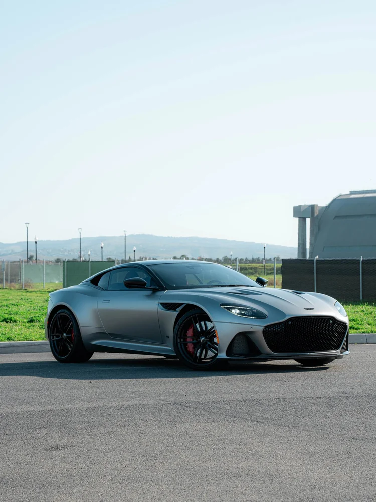Matte silver-grey Aston Martin DBS in a complete front three-quarter view with black wheels and red brake calipers outdoors