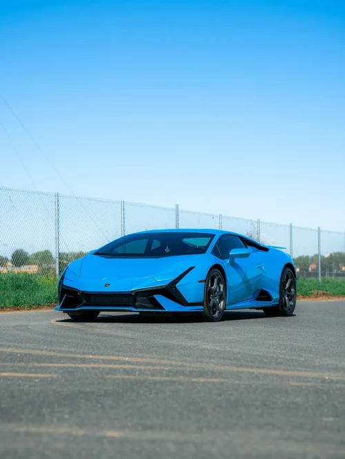 Blue Lamborghini Huracán Tecnica in a front three-quarter view on a road beside grass and a chain-link fence