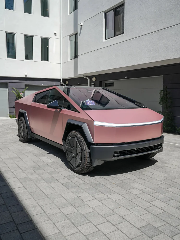 Pink-gold Tesla Cybertruck in a complete front-three-quarter view with dark angular wheel covers on courtyard paving.
