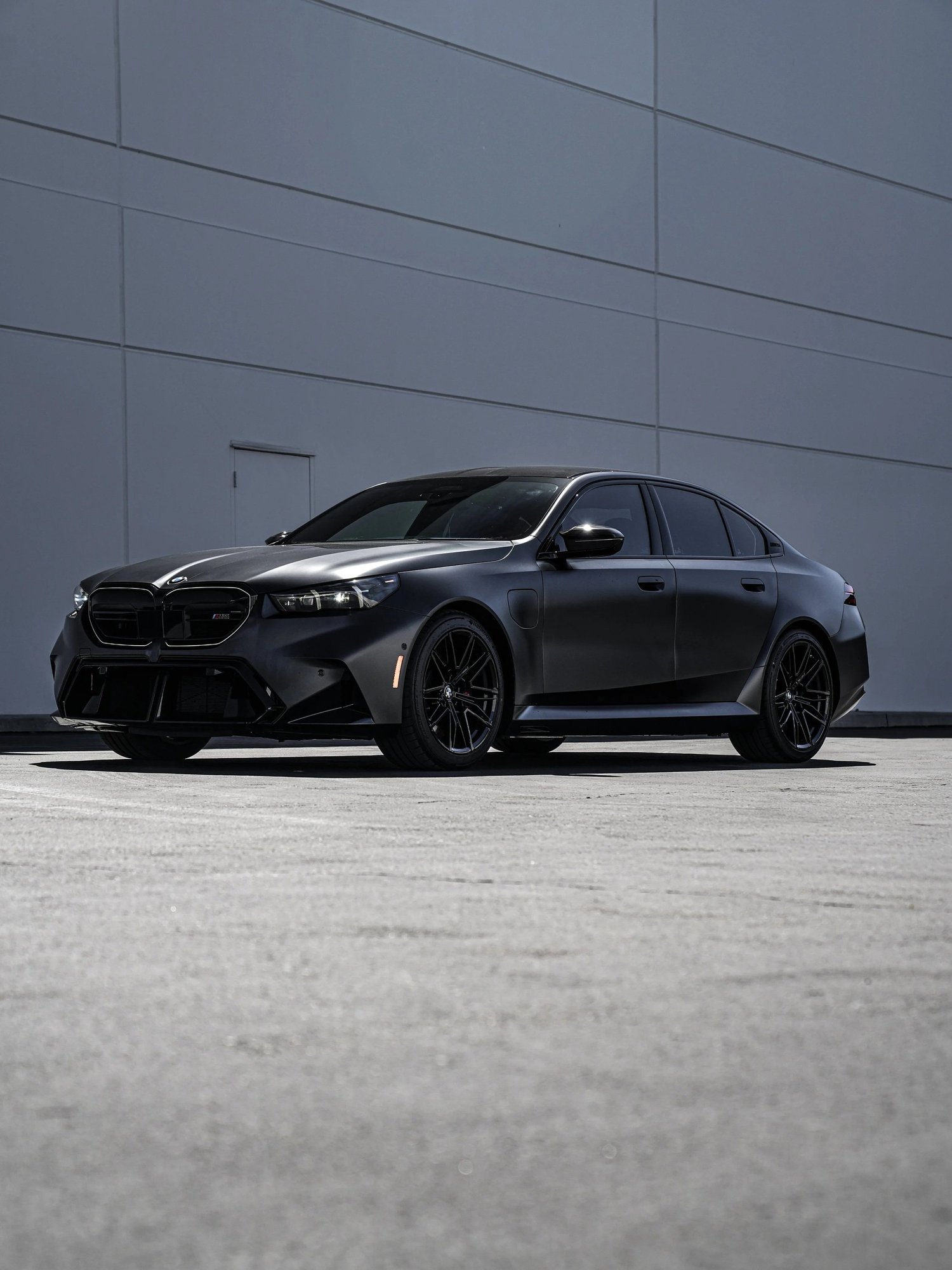 BMW G90 M5 in a low front three-quarter view, with dark matte body panels, black wheels and side glass visible