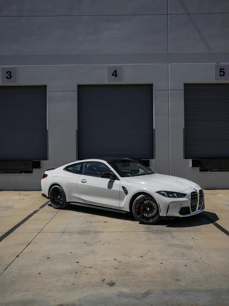 Complete front-three-quarter view of a white BMW G82 M4 coupe with dark wheels and a dark roof outside numbered garage doors.