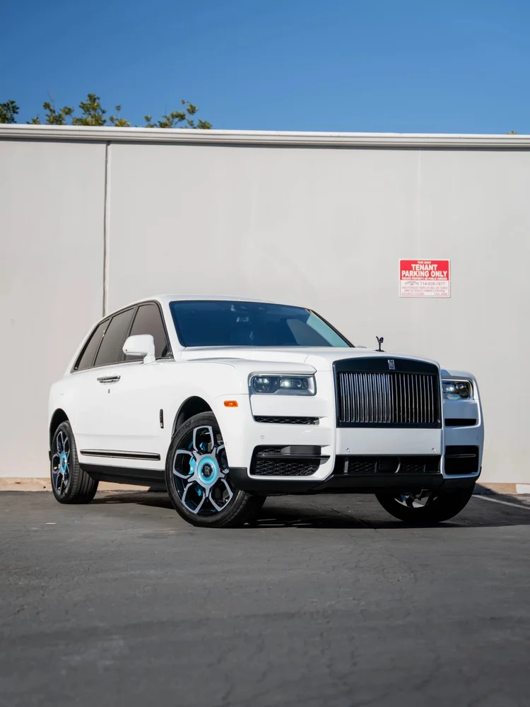 Complete front three-quarter view of a white Rolls-Royce Cullinan with a dark grille, turquoise wheel accents and dark side glass beside a pale workshop wall.