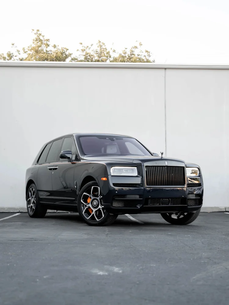 Black Rolls-Royce Cullinan Black Badge, complete front-three-quarter view with orange brake calipers beside a light wall