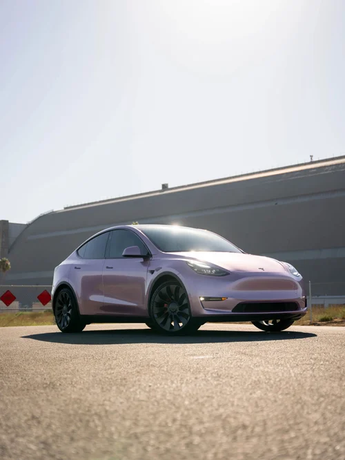 Pale pink Tesla Model Y in a front three-quarter view in a sunlit paved lot.