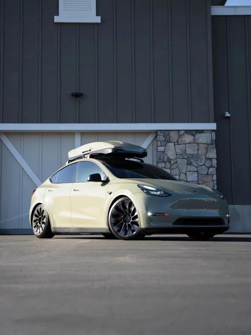 Khaki green Tesla Model Y with a roof cargo box in a front three-quarter view beside a building.