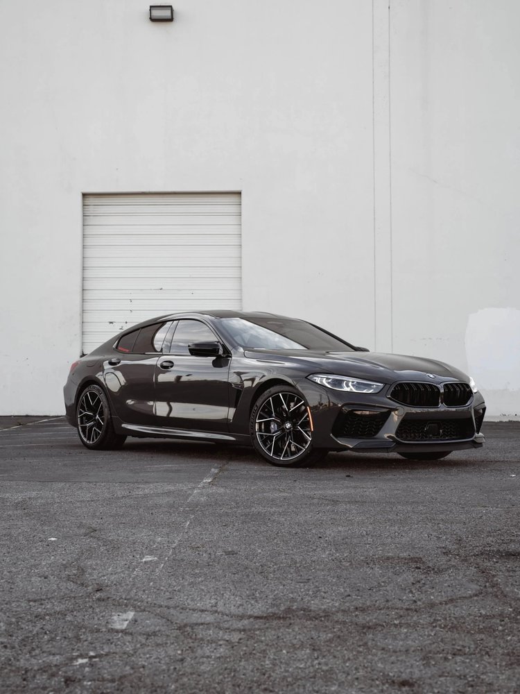 Complete front three-quarter view of a reflective dark BMW M8 with black-and-silver wheels outside a pale industrial wall and roll-up door.