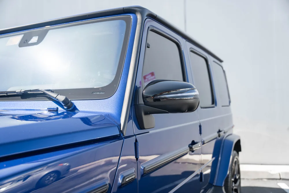 Reflective blue G 63 body beside the black mirror, windshield and dark side windows in a close side view