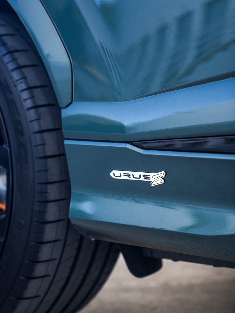 Close side-skirt detail of the green Lamborghini Urus S showing a silver Urus S badge, black trim and the edge of a tire.