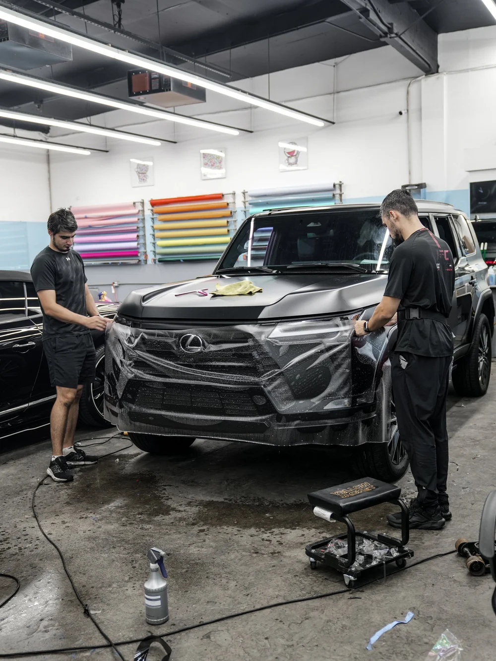 Two installers holding a broad sheet of wet transparent film across the Lexus front bodywork beneath workshop lights, with a yellow cloth on the hood