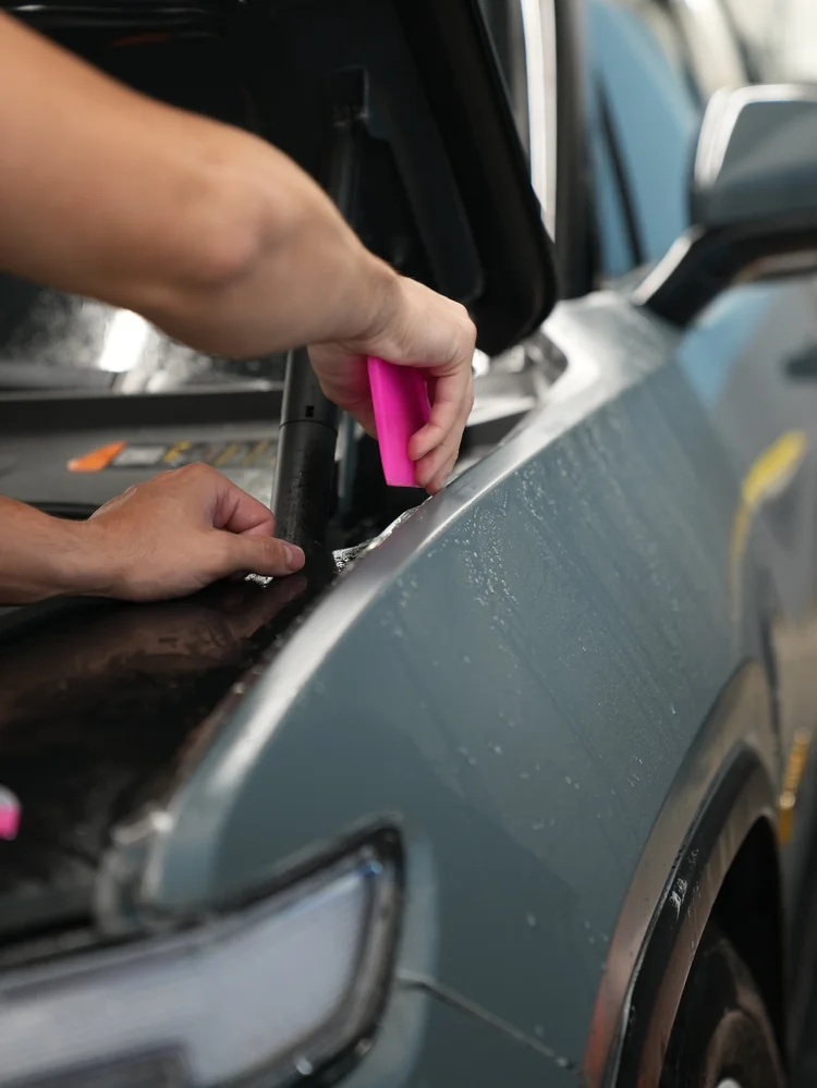 Hands working a pink squeegee beside wet film at a Rivian R1T front-panel edge with the hood open.