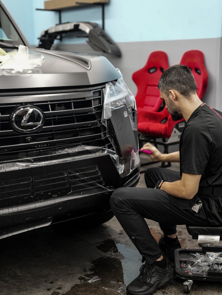 Seated installer using a pink tool beside transparent film at the Lexus front corner, with wet film across the grille and two red seats in the background