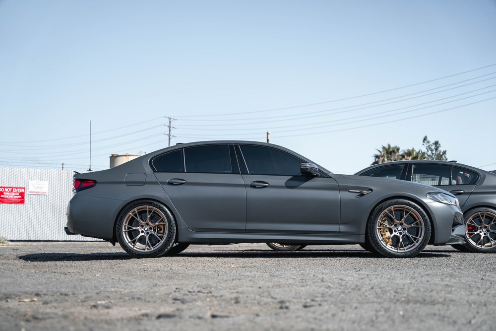 Gray BMW M5 CS in complete side profile with bronze-colored wheels and dark side windows, with its companion behind