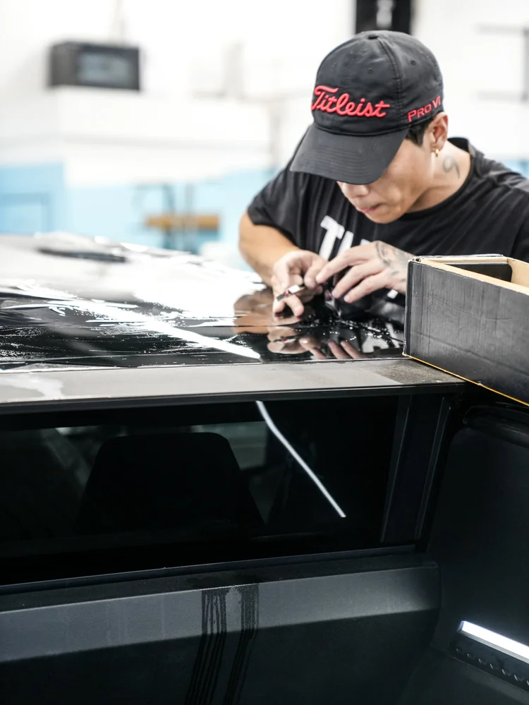 A technician using a small cutting tool near wet film at the upper Tesla Cybertruck panel edge, with a film box nearby.