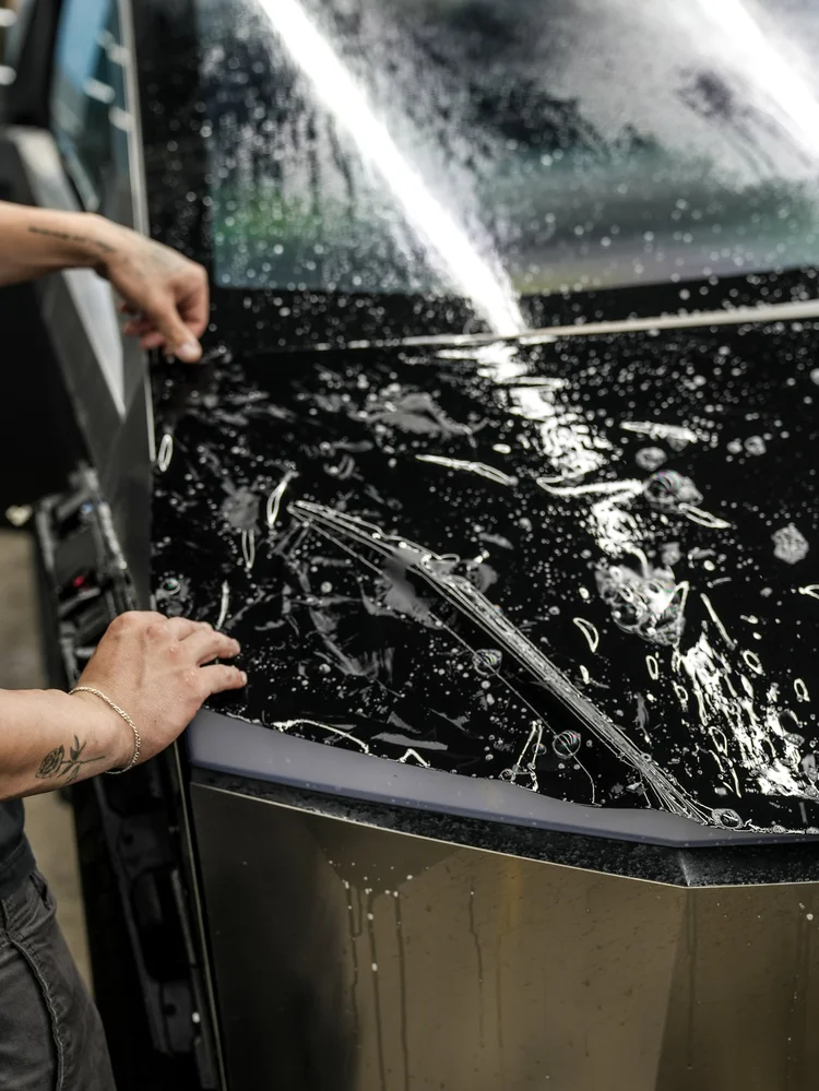 Hands holding wet black film over an angular Tesla Cybertruck body panel during installation, with droplets and stretched film visible.