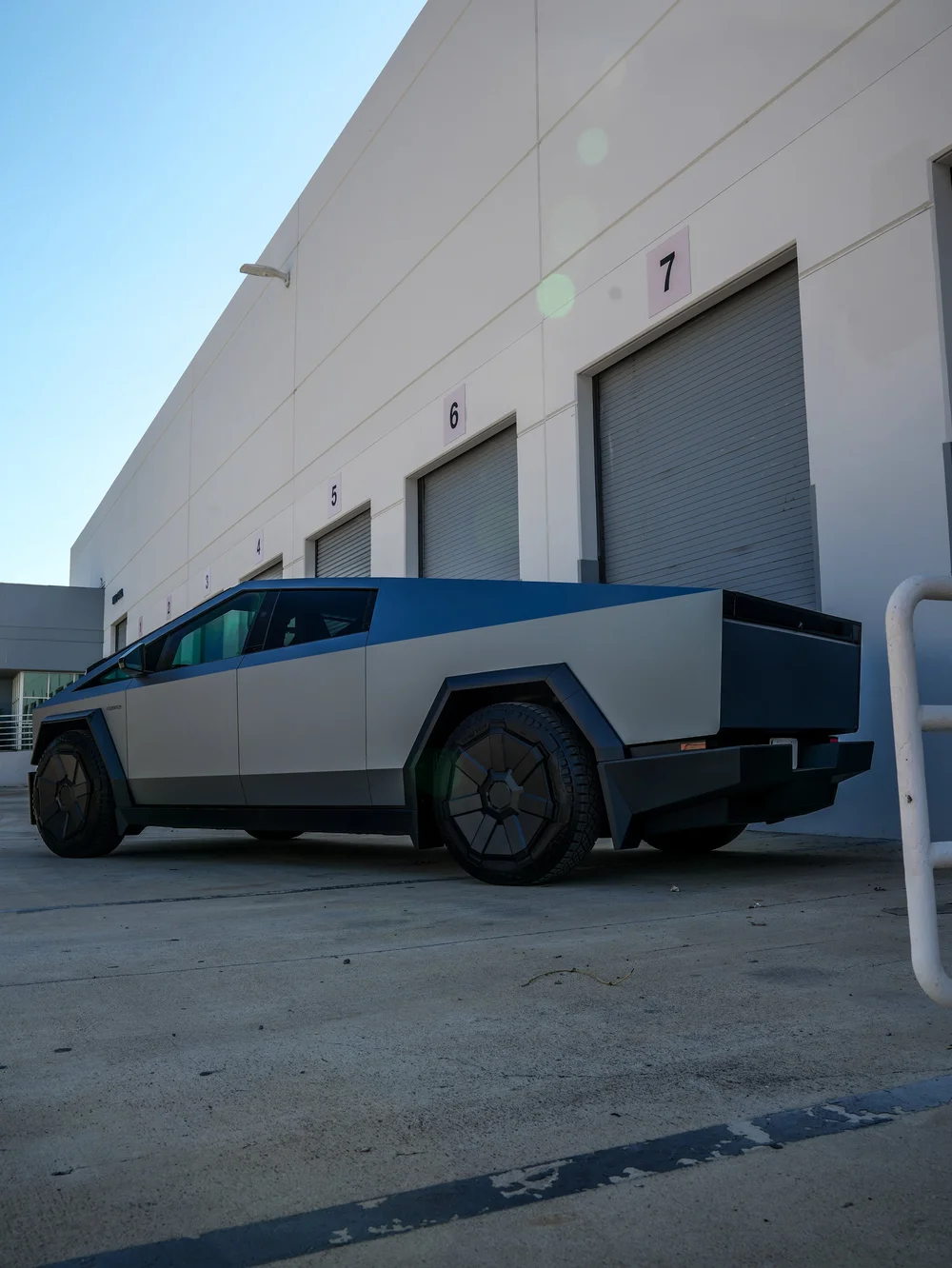 Complete low left-facing rear three-quarter view of the grey blue Tesla Cybertruck with its long side panels and dark geometric wheel covers beside numbered doors.