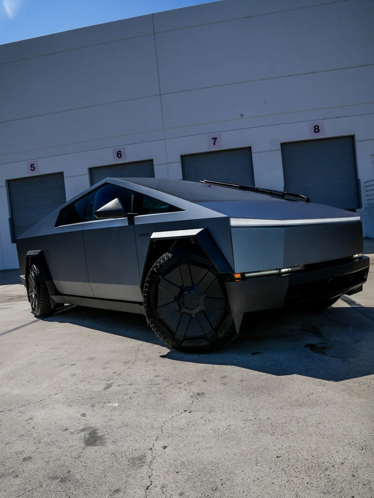 Low right-facing front three-quarter view of the grey blue Tesla Cybertruck with dark wheel covers and angular front bodywork beside numbered workshop doors.