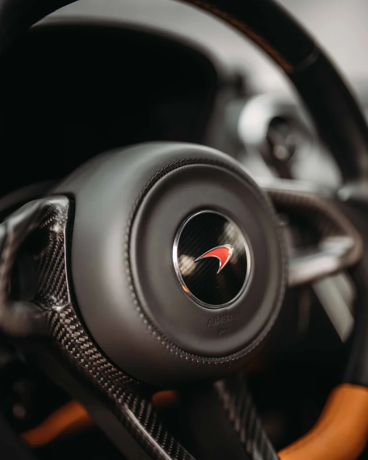 Close steering-wheel view of the red McLaren emblem, stitched center and patterned spokes.