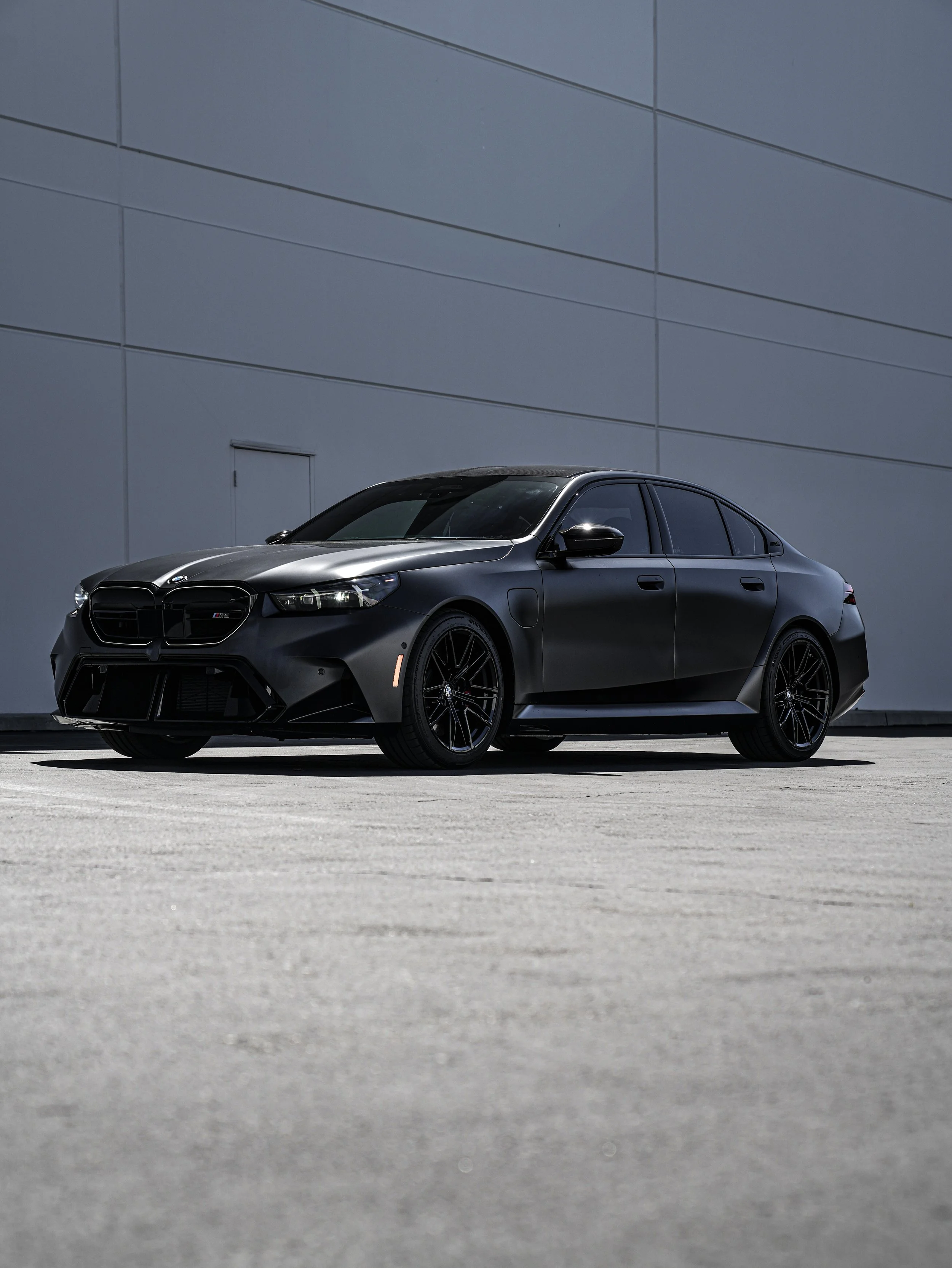 A matte black BMW sedan parked on a concrete surface against a gray wall.