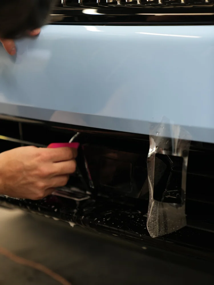 Clear film hanging over the Cullinan front bumper beside a dark lower intake as a technician uses a pink tool